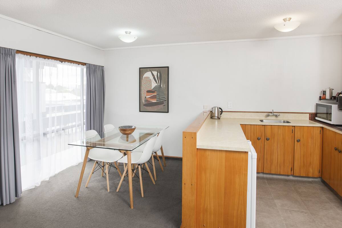 A modern kitchen with wooden cabinets, a glass dining table, and a large window with curtains.