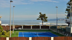 A tennis court with a wooden fence, outdoor seating, and a view of the ocean. - Waikanae Beach Motel (Gisborne)