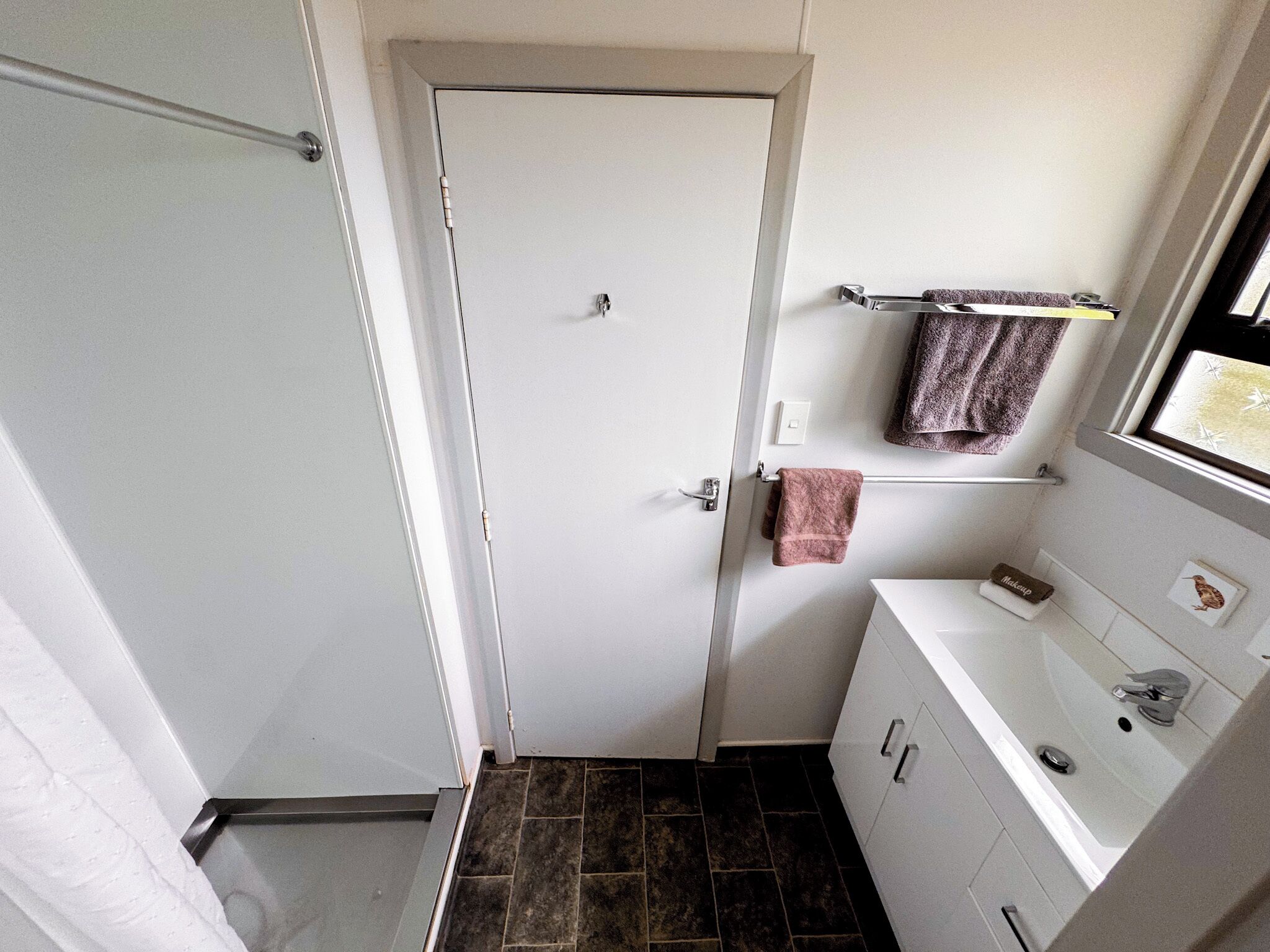 A small bathroom with a white door, a towel rack with two towels, a white sink with a mirror, and a window above the sink.