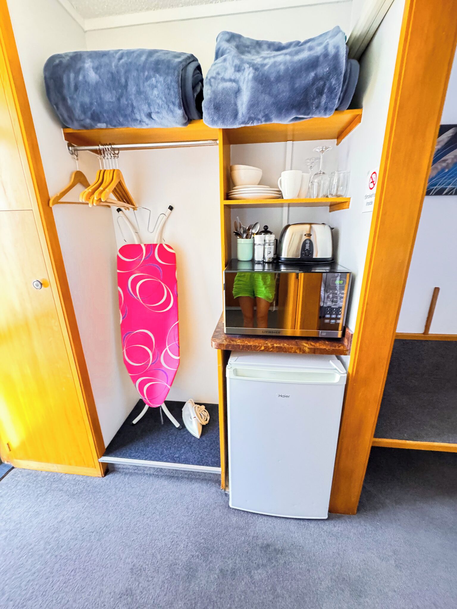 A compact room with a bed, a small refrigerator, a microwave, a toaster, and a shelf with dishes and glasses.