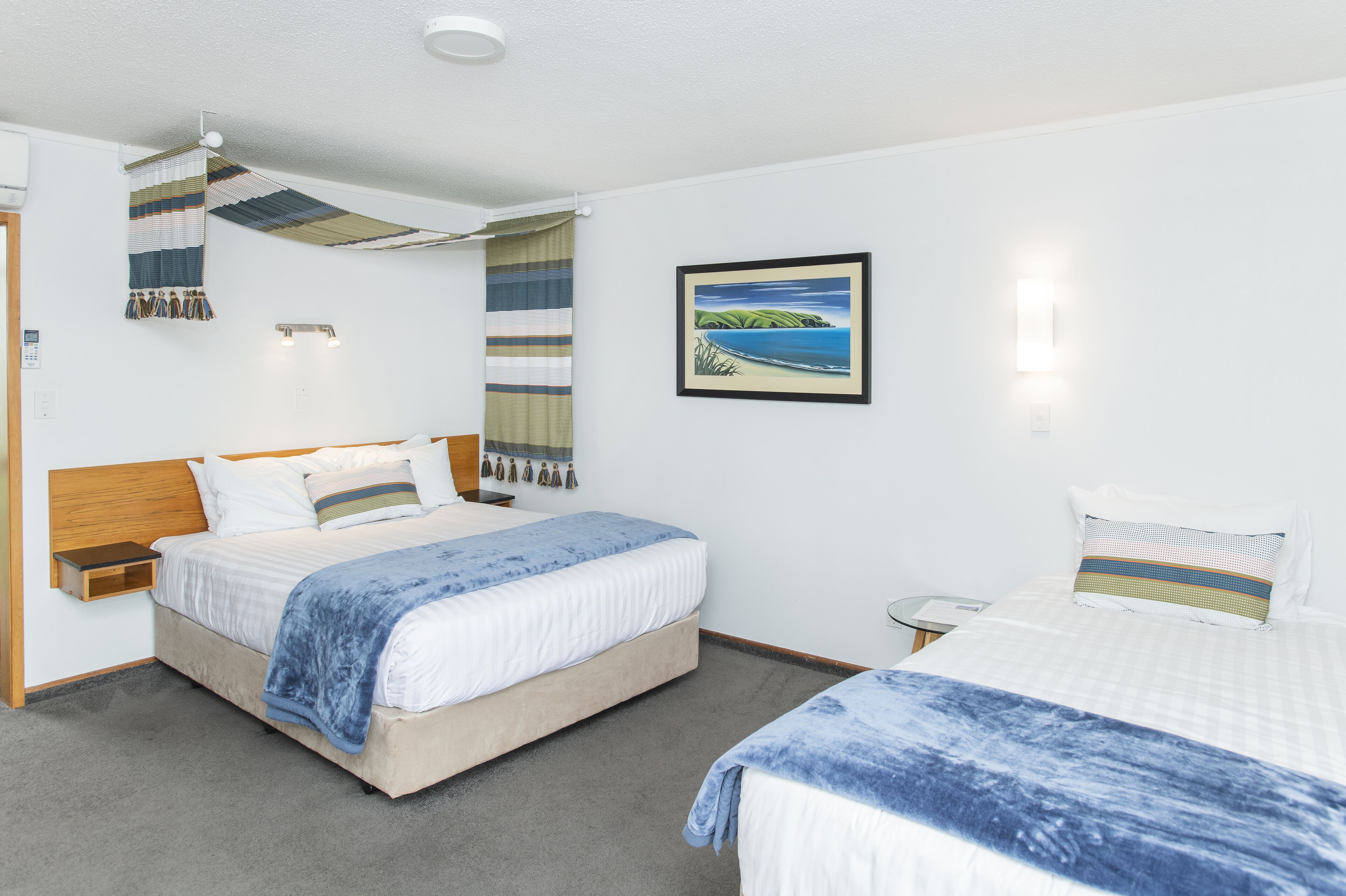 A hotel room with a bed, a bedside table, a nightstand, a wall-mounted lamp, a framed picture, and a hanging tapestry.