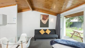 Una habitación con una mesa de vidrio, dos sillas blancas, un sofá gris con dos cojines amarillos, una cama con una colcha azul y un techo de madera.