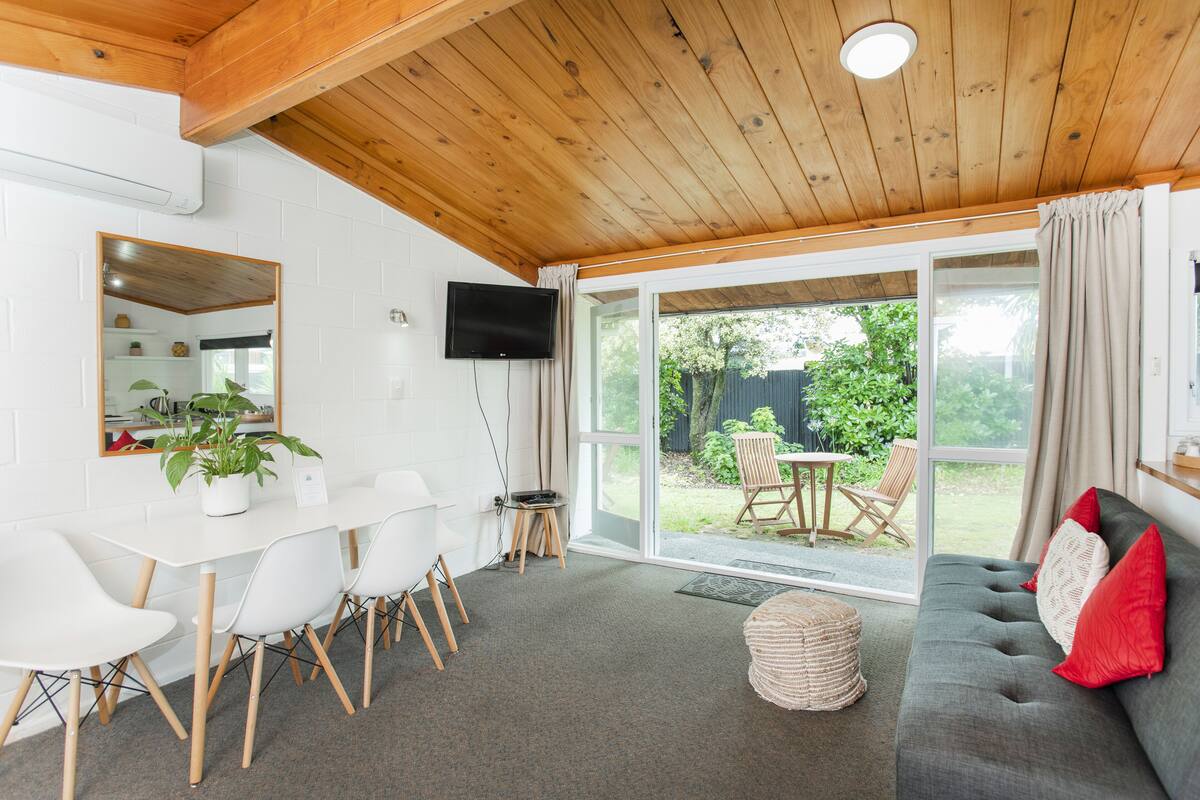 A modern living room with a wooden ceiling, a grey sofa with red cushions, a white dining table with chairs, and a large sliding glass door leading to an outdoor area.