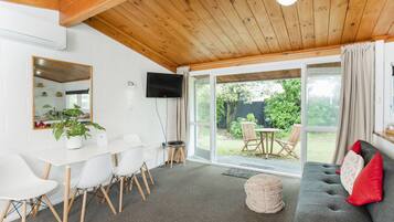 Una sala de estar moderna con techo de madera, un sofá gris con cojines rojos, una mesa de comedor blanca con sillas y un amplio ventanal corredero que da a un espacio exterior.