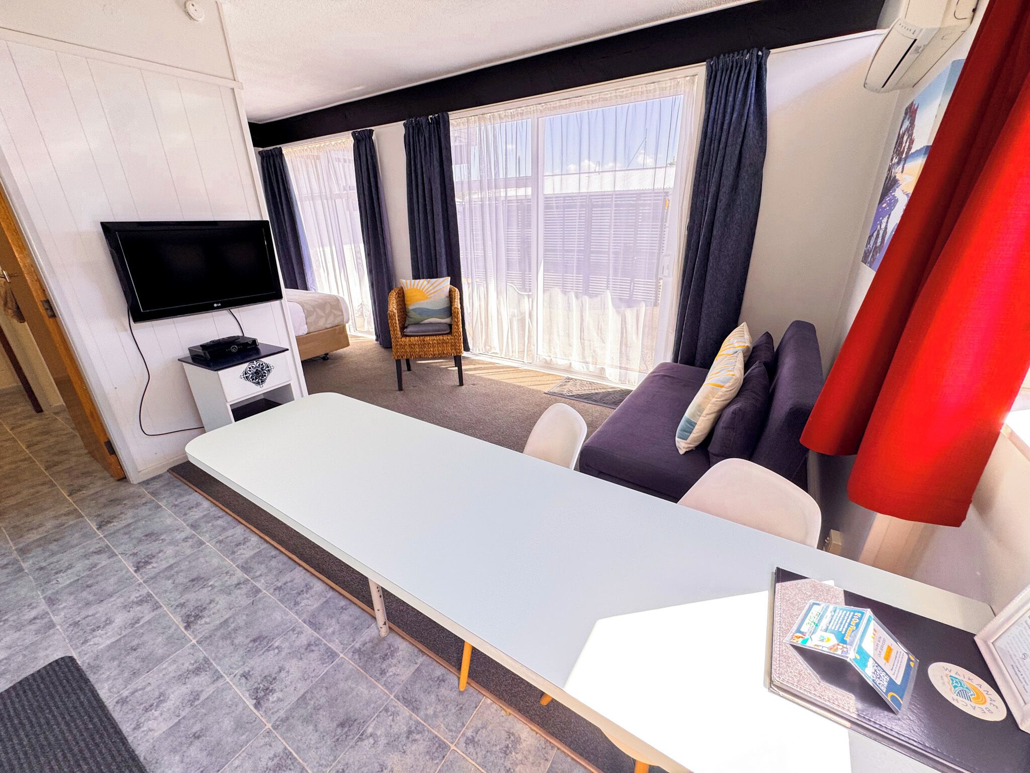 Una habitación de hotel moderna con televisión de pantalla plana, una mesa de comedor, un sofá y una ventana con cortinas.