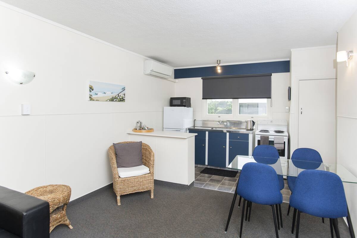 A compact kitchen with a microwave, refrigerator, and stove, a dining area with blue chairs, a wicker chair, and a small table.