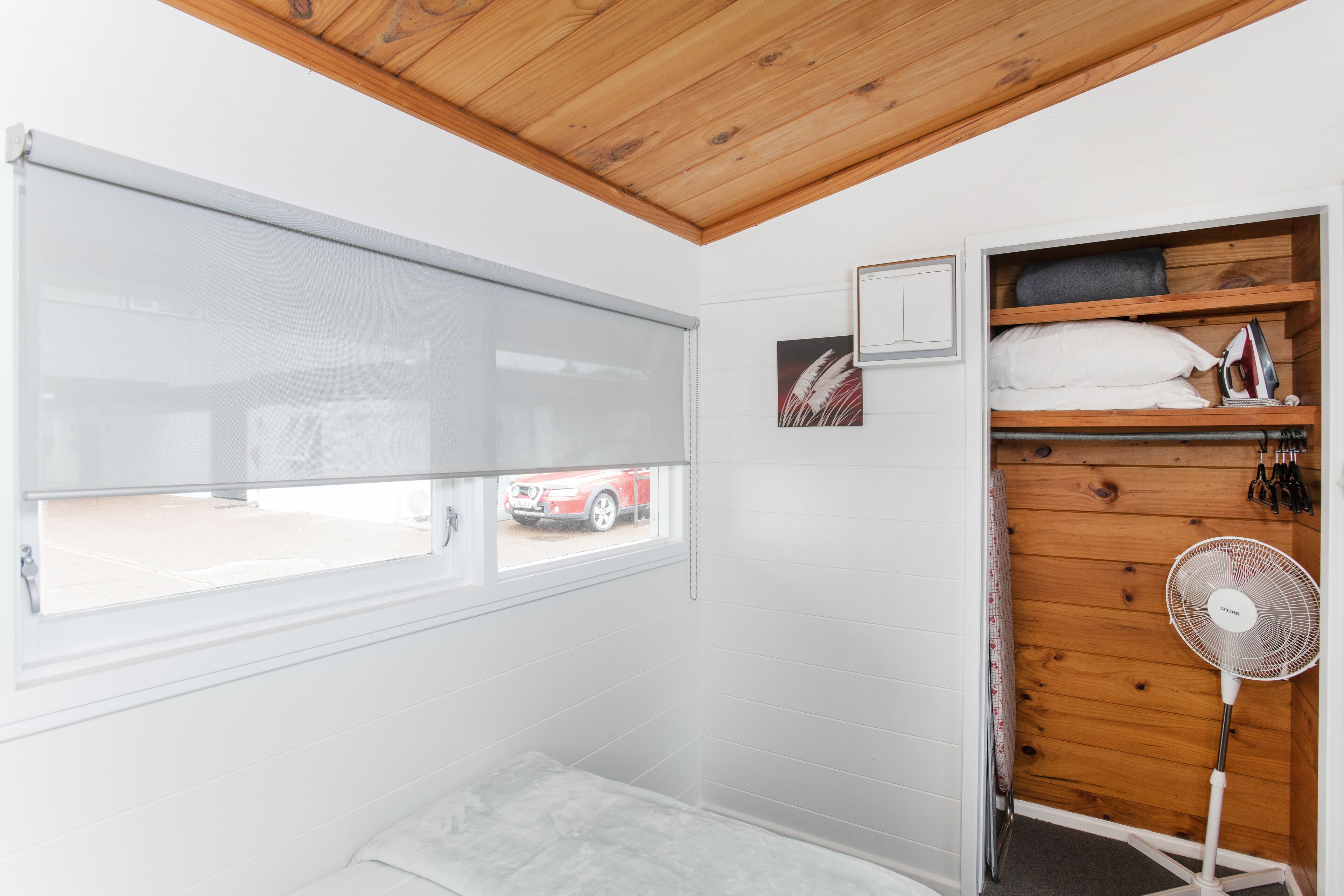 Um quarto com uma janela grande, teto de madeira, uma cama, um ventilador e uma estante com toalhas e um ferro de passar roupa.