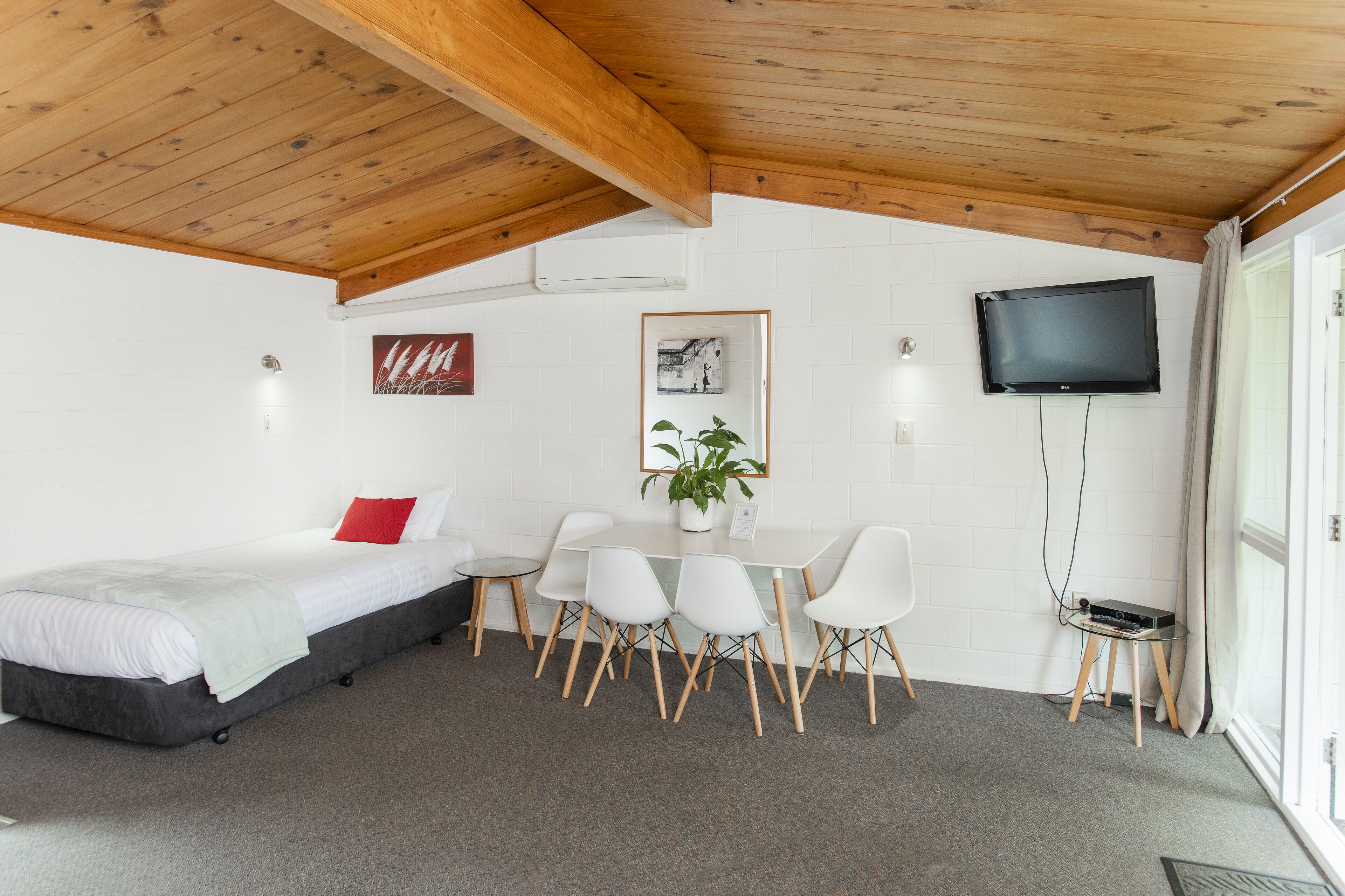 A bedroom with a bed, a desk, chairs, a TV, and a plant.