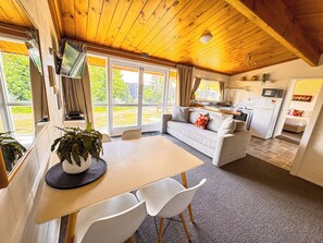 Una habitación acogedora con techo de madera, una mesa de comedor con sillas, un sofá, una zona de cocina y ventanas amplias.