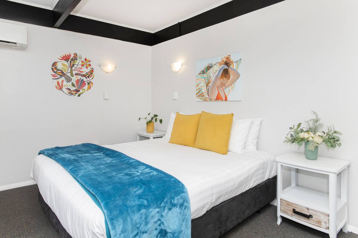 A bedroom with a bed, white walls, a blue blanket, yellow pillows, a nightstand with a vase of flowers, and two wall art pieces.