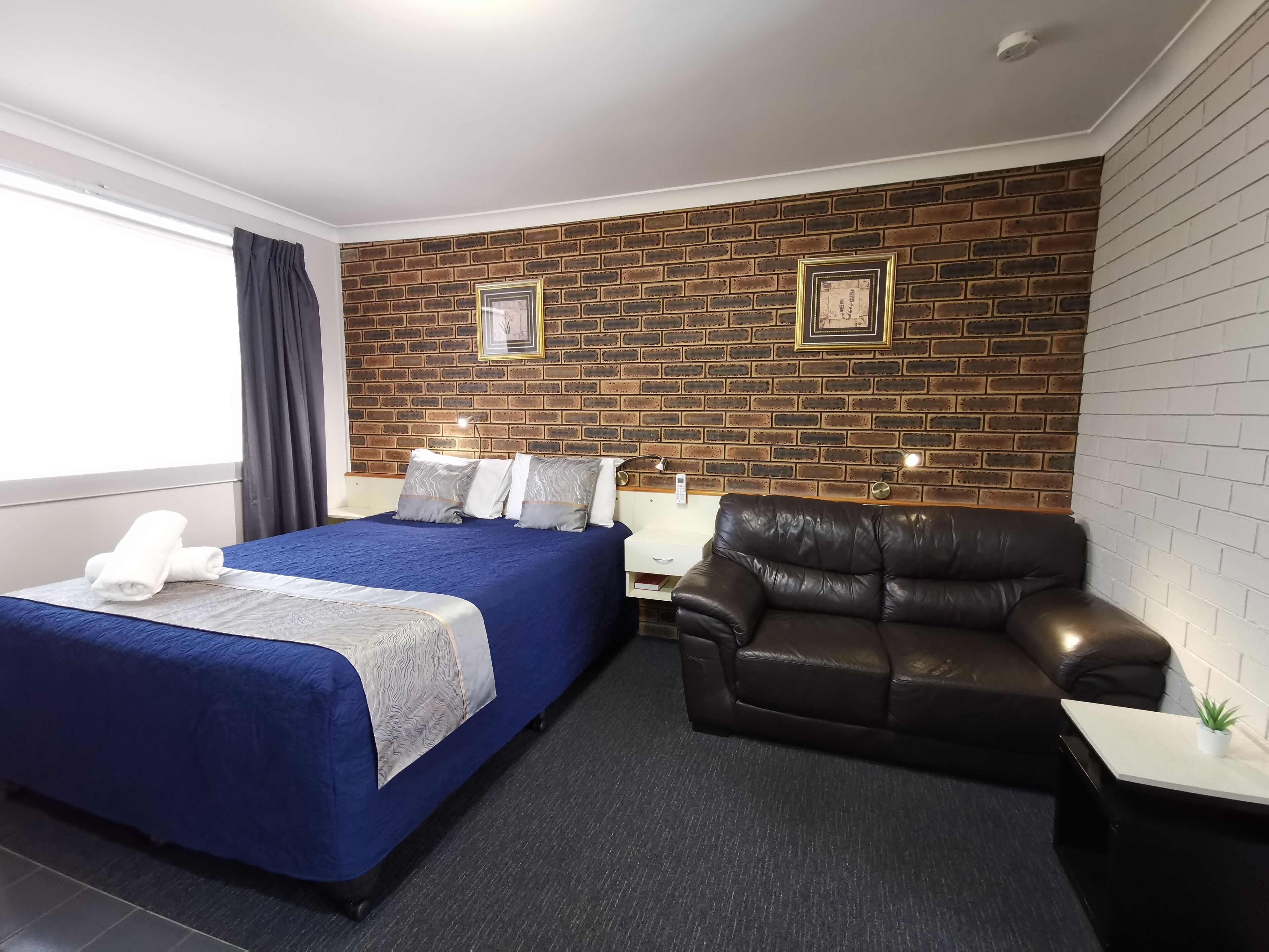 Habitación de hotel con pared de ladrillo, cama con cubrecama azul, sofá de cuero marrón y una mesita con planta.