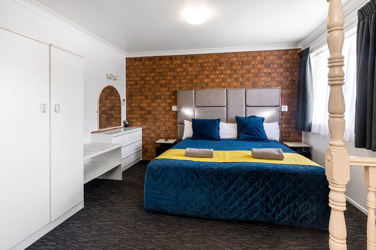 A hotel room with a brick wall, a large bed with blue bedding, a wooden headboard, a white dresser, and a wooden nightstand.