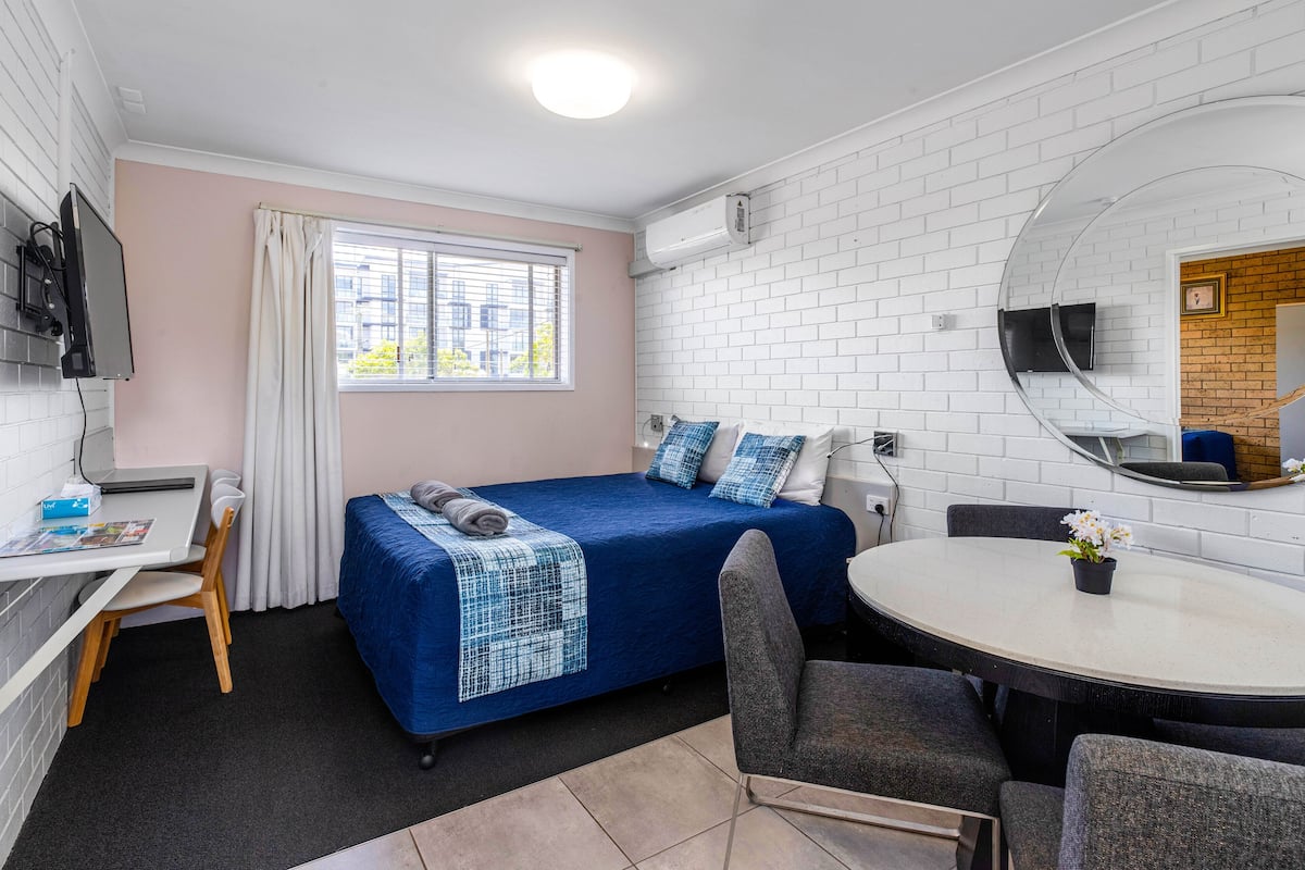 A compact hotel room with a single bed, a desk, a chair, a round dining table, and a TV mounted on the wall.