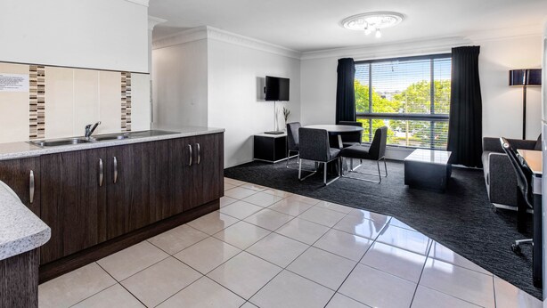 A modern hotel room with a kitchen area, a dining table with chairs, a flat-screen TV mounted on the wall, and a large window with curtains. - Leichhardt Motor Inn (South Toowoomba)