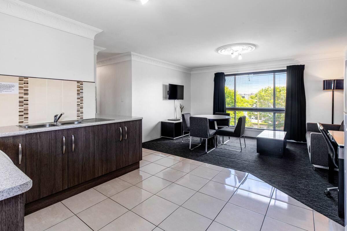 A modern hotel room with a kitchen area, a dining table with chairs, a flat-screen TV mounted on the wall, and a large window with curtains.