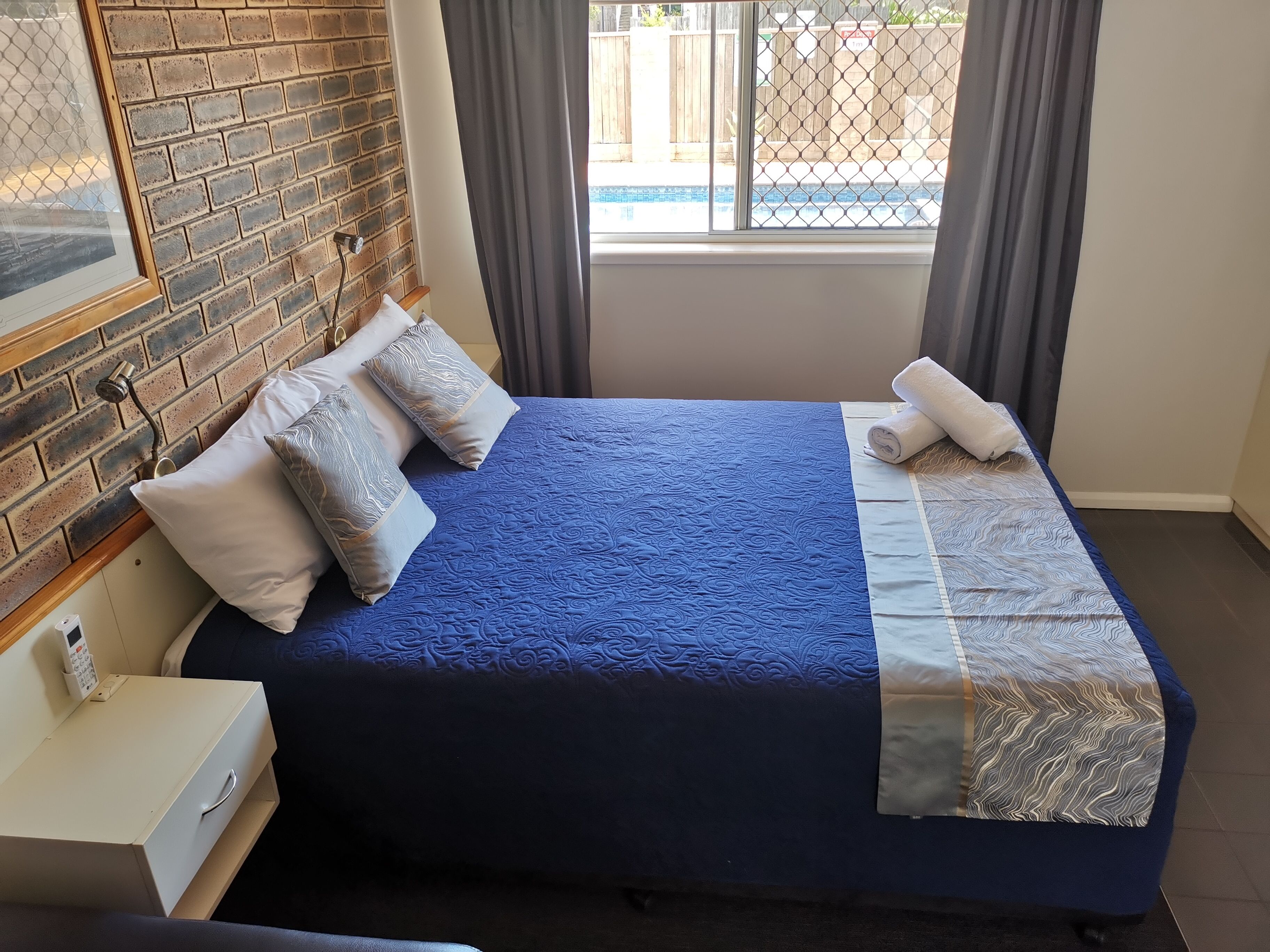 A neatly made bed with a blue quilt, a white pillow, and a patterned pillow. There is a small bedside table with a remote control and a white box. A window with a view of a pool and a fence is visible.