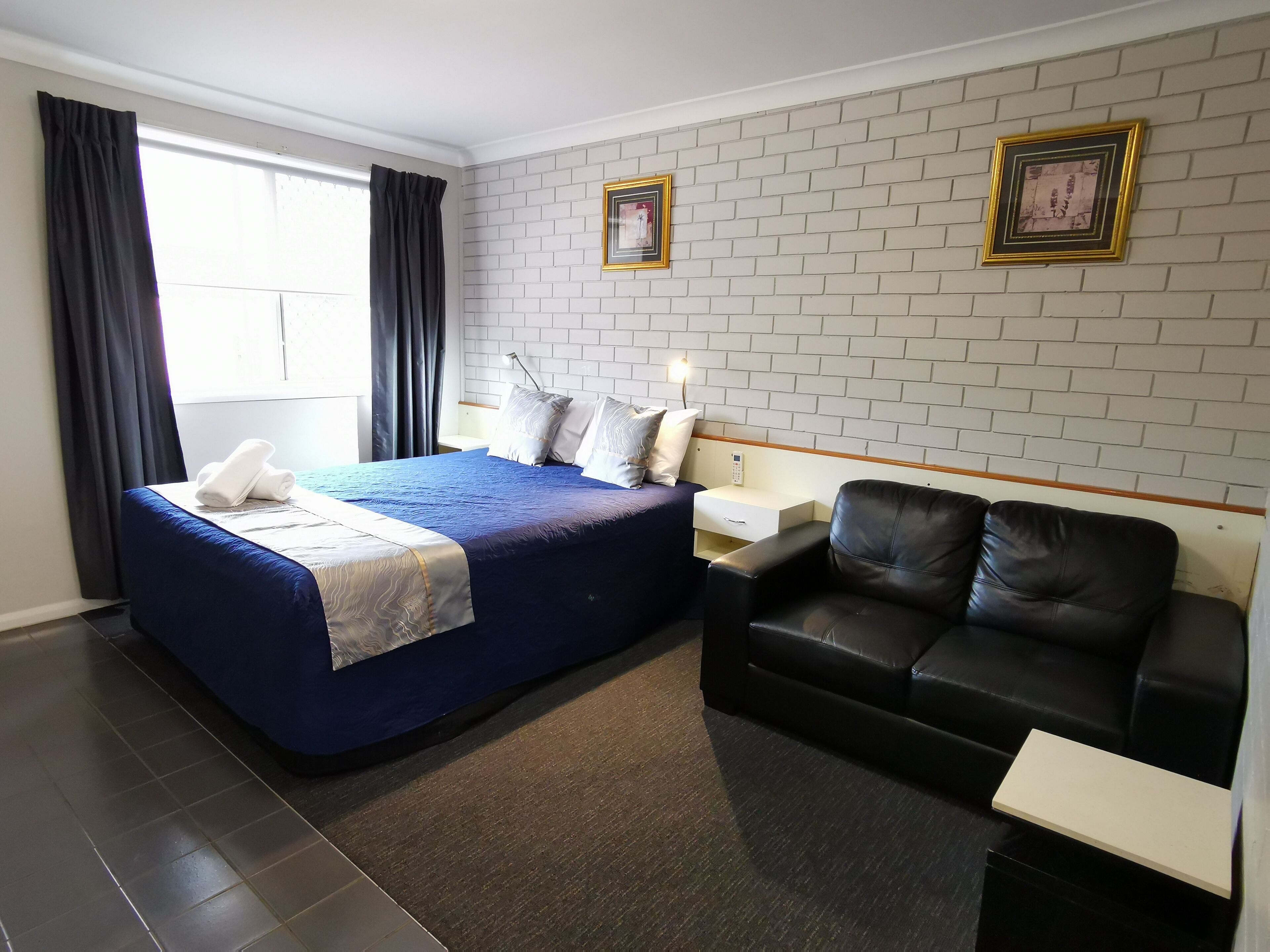 Habitación de hotel con una cama, un sofá de cuero negro, una mesita de noche y una pared con dos cuadros enmarcados.