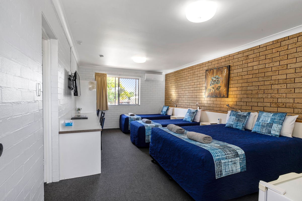 A hotel room with three beds, a white brick wall, a wooden accent wall, and a window with curtains.