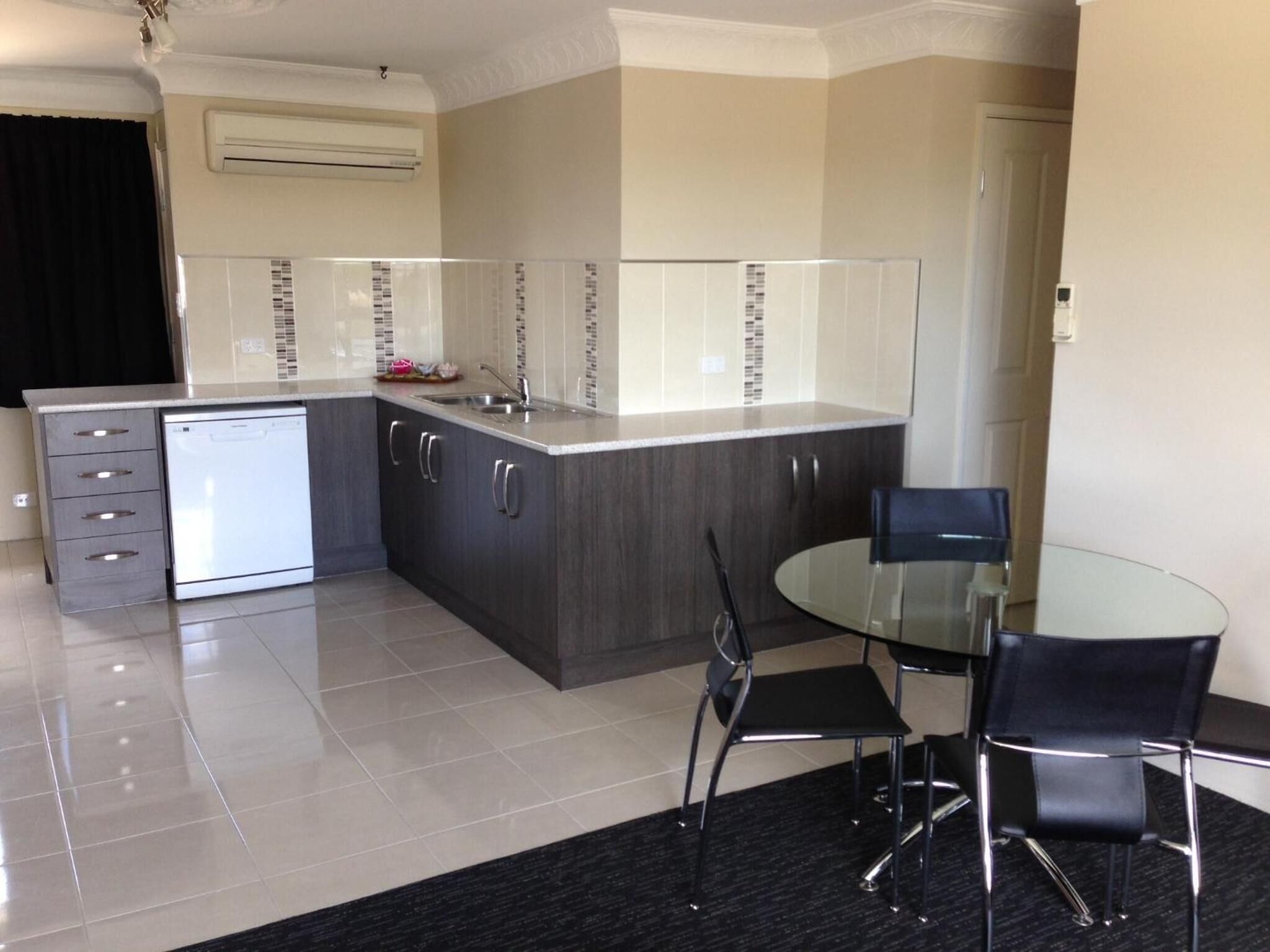 A modern kitchen with a dishwasher, dark cabinets, a glass dining table, and two chairs.