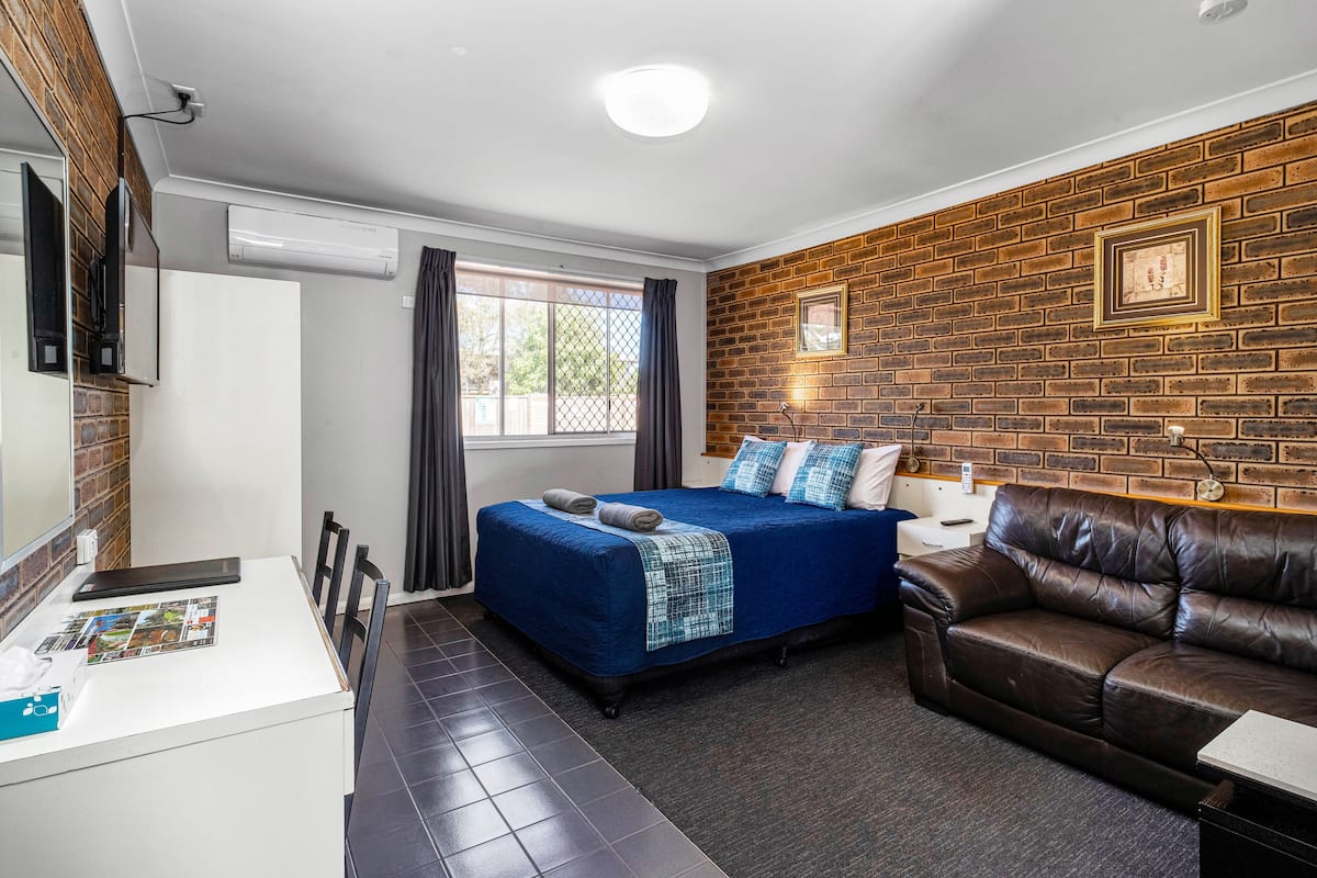 A modern room with a brick wall, a bed with blue bedding, a leather sofa, a desk with a tablet, and a window with curtains.