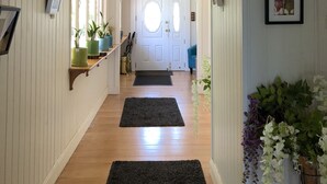 Un pasillo con pisos de madera, paredes blancas y una puerta blanca al final. Hay plantas en macetas y cuadros en las paredes.