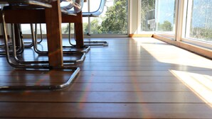 Suelo de madera con sillas y una mesa, ventanas grandes con vistas a árboles.