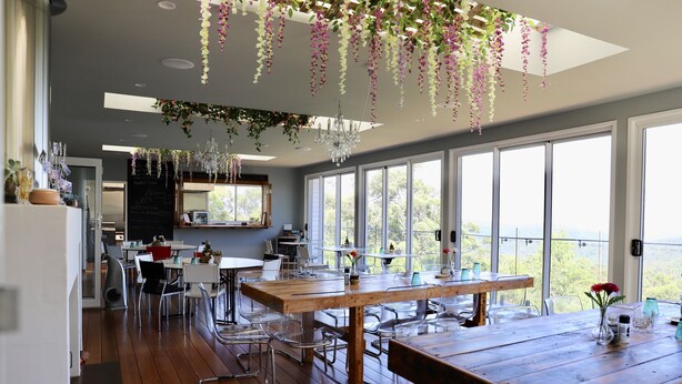 Un comedor espacioso con una mesa y sillas de madera, ventanas amplias que ofrecen vistas al exterior y decoración floral colgante.