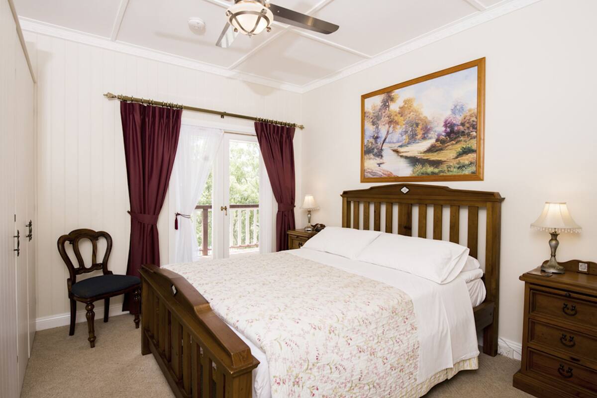 Ein Schlafzimmer mit einem hölzernen Bett, einem Sessel, einem Nachttisch, einem Gemälde und einem Fenster mit Vorhängen.