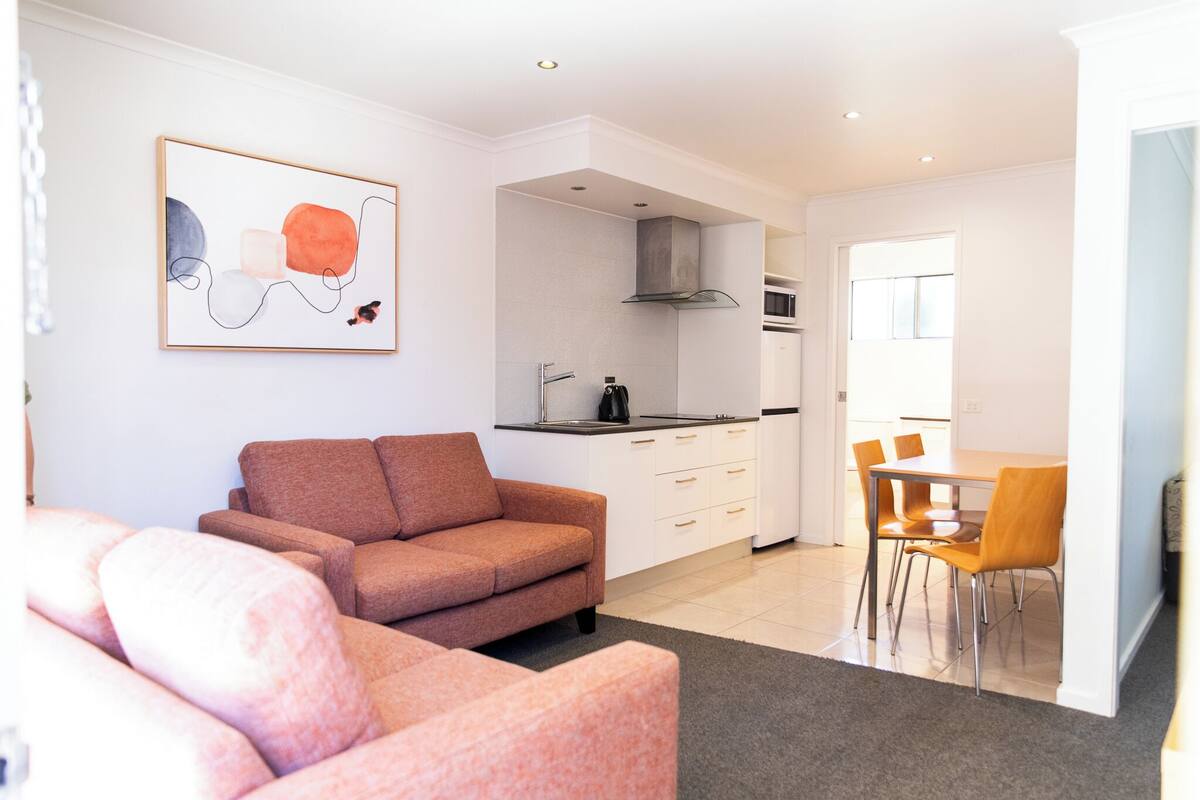 A modern living room with a sofa, a dining area with chairs, a kitchen with a stove and microwave, and a painting on the wall.