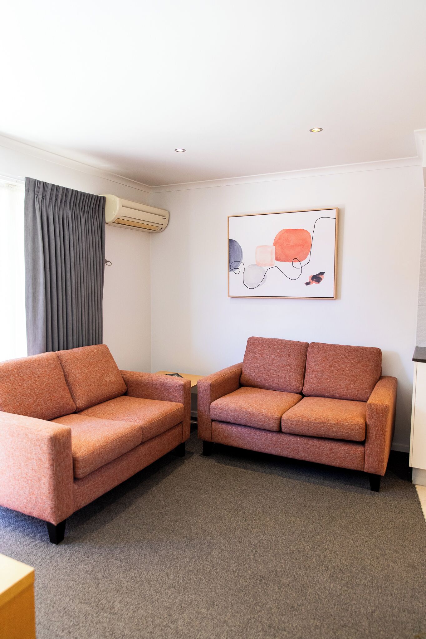 A room with two orange sofas, a grey carpet, a wall-mounted air conditioner, and a framed abstract painting.