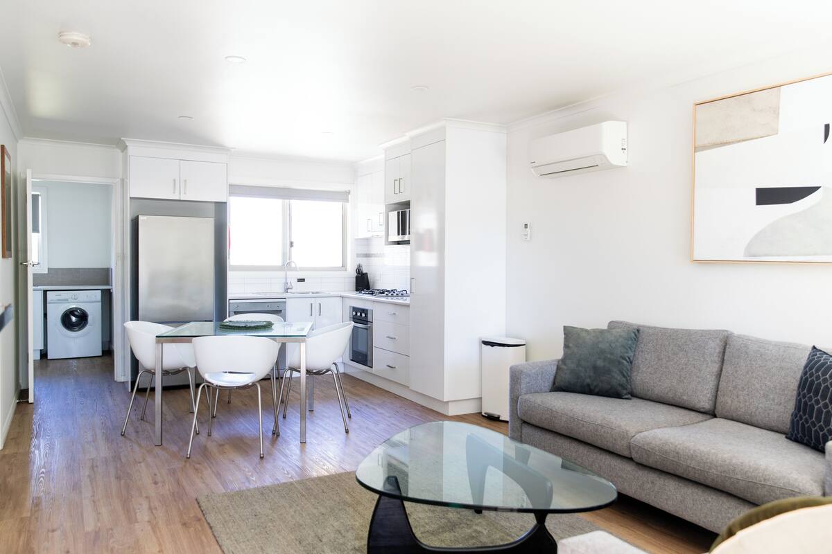 A modern kitchen with white cabinets, a dining area with a glass table, and a grey sofa.