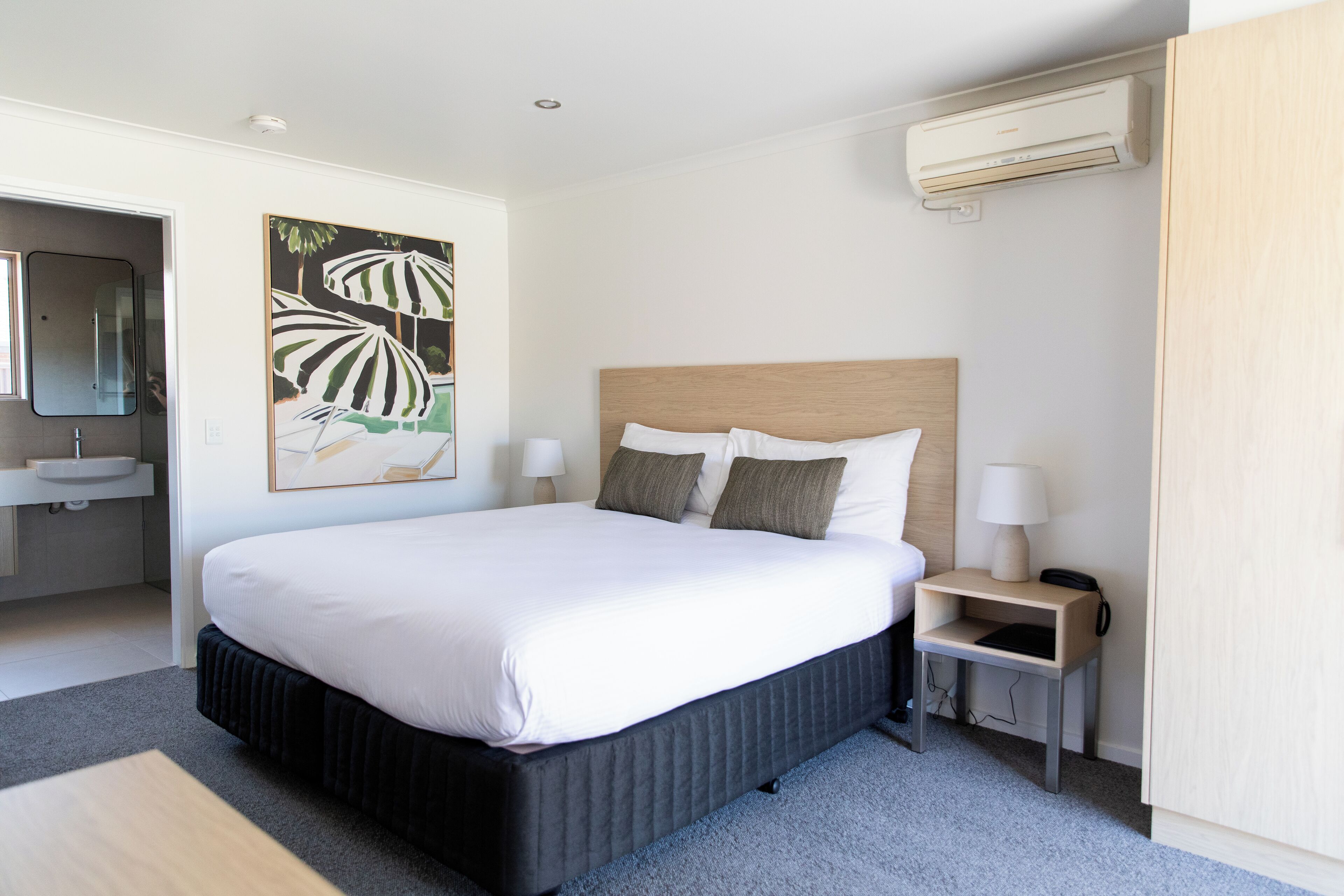 Une chambre d’hôtel moderne avec un grand lit, des tables de chevet avec des lampes, une salle de bain avec un lavabo et un tableau au mur.