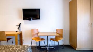 A hotel room with a bed, a desk with a chair, a television mounted on the wall, and a wardrobe.