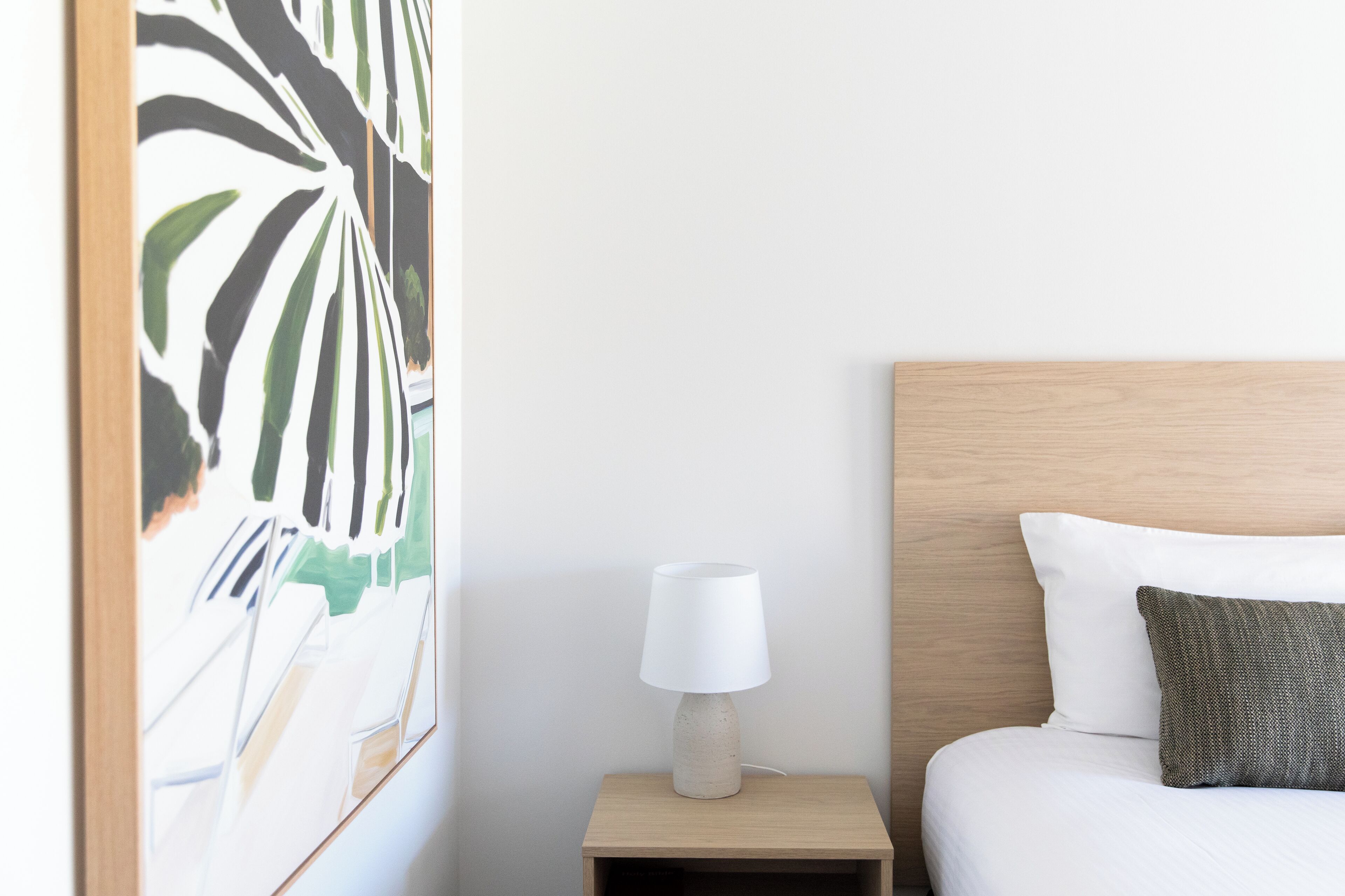 Una cama bien tendida con ropa de cama blanca, un cabecero de madera y una mesita de noche con una lámpara blanca.
