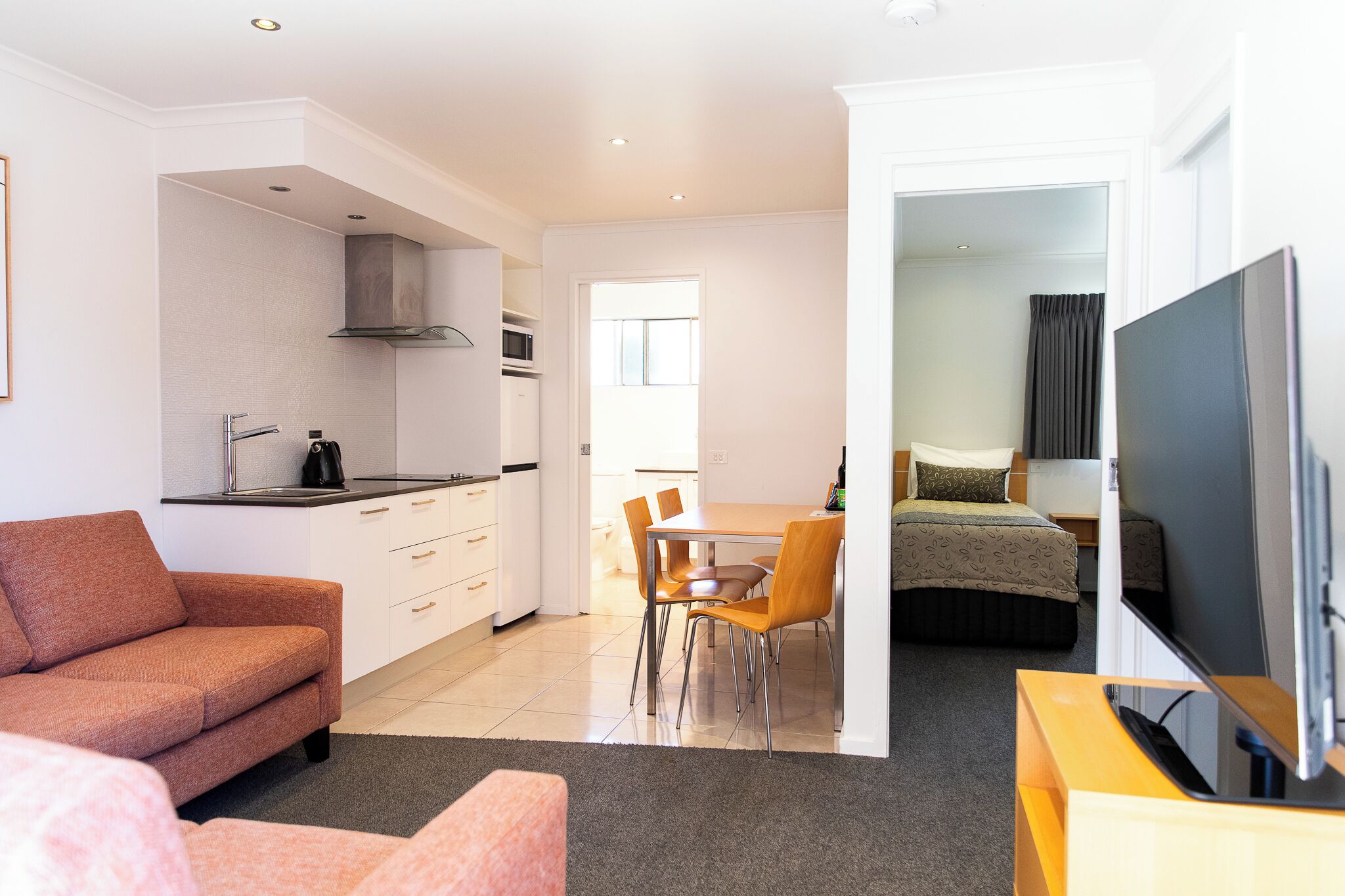 A modern hotel room with a kitchenette, a dining area, a sofa, and a TV.