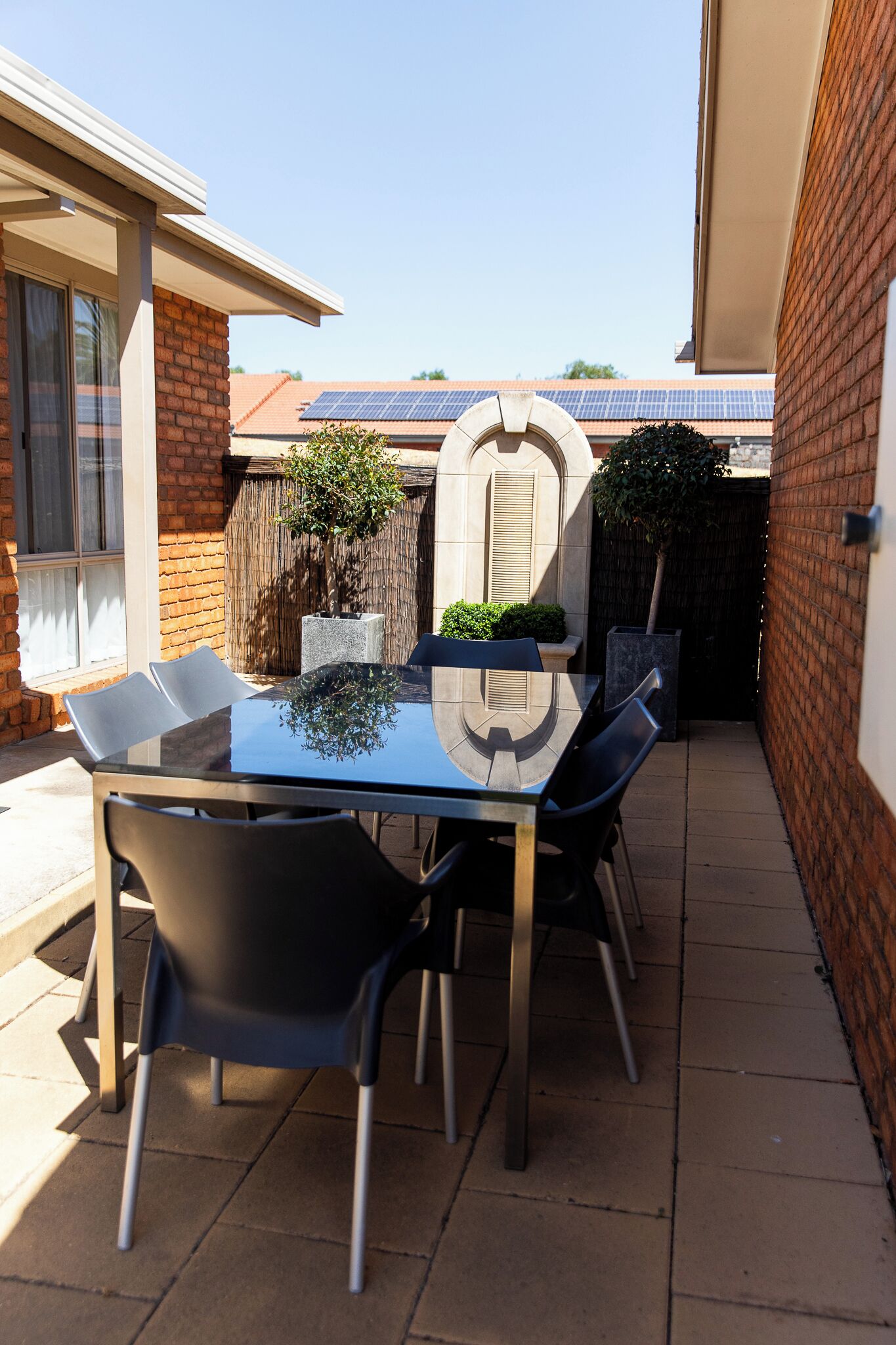 Un patio con una mesa y sillas de vidrio, rodeado de muros de ladrillo y un pequeño árbol.