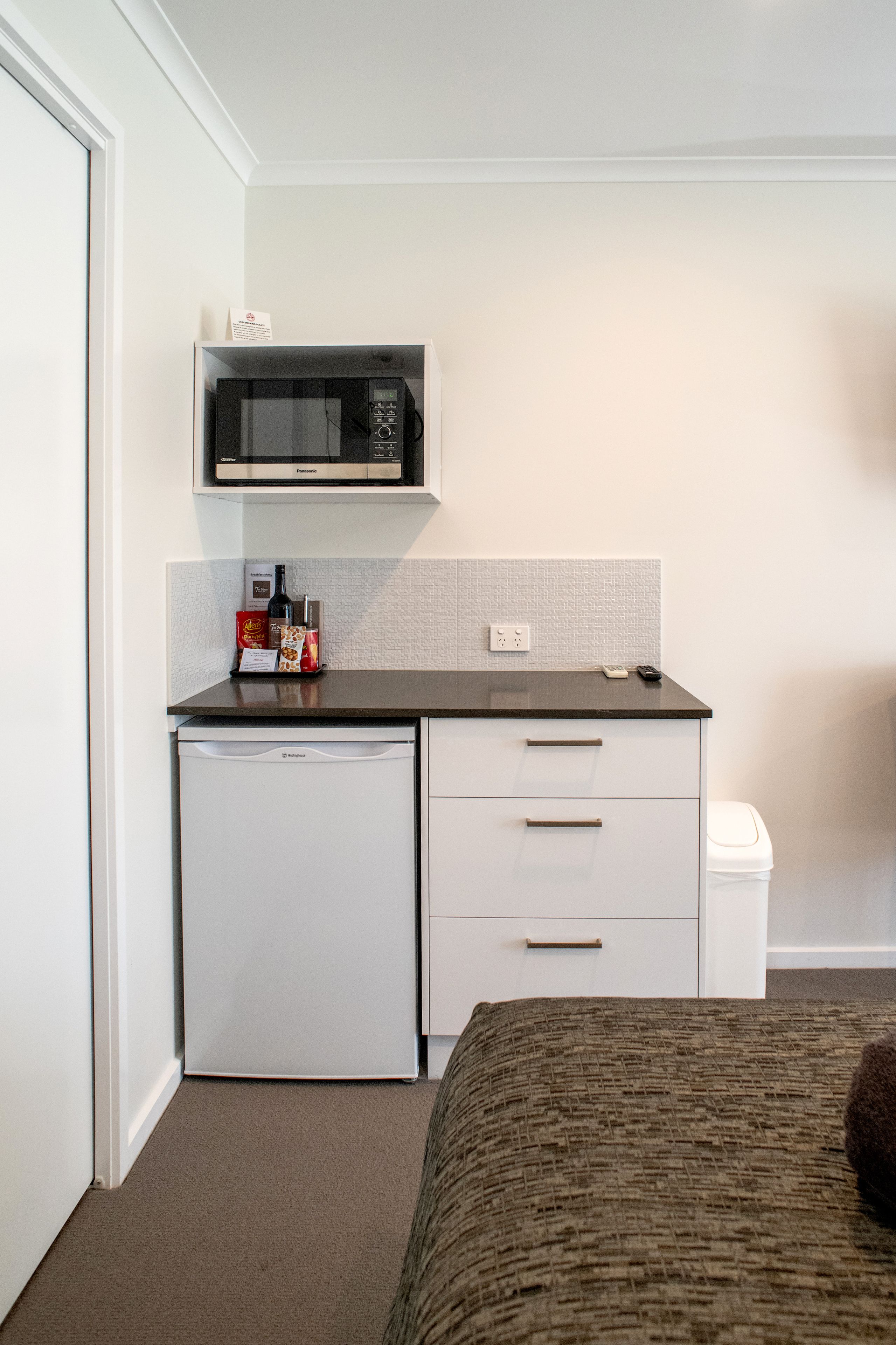 Uma kitchenette compacta com micro-ondas, um frigorífico pequeno e uma máquina de café. Há uma cama com um cobertor castanho e um travesseiro.