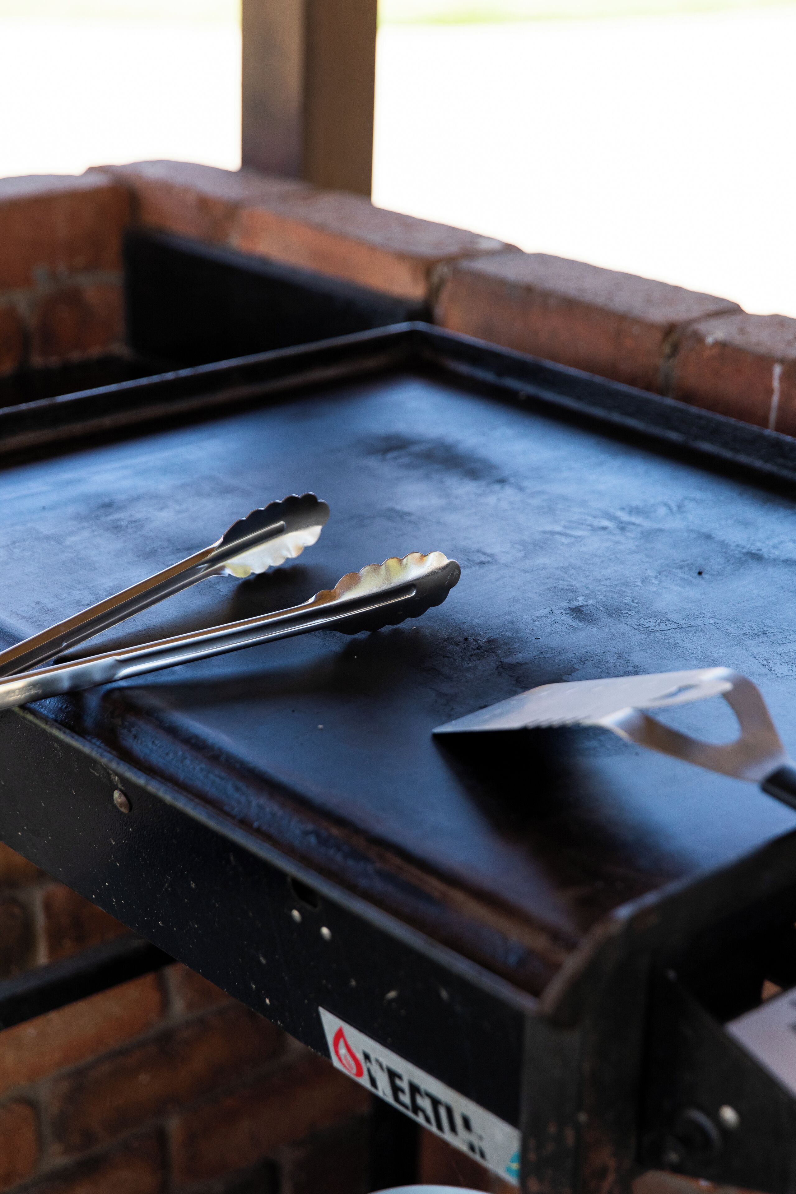A metal tray with tongs and a spatula on a brick surface.