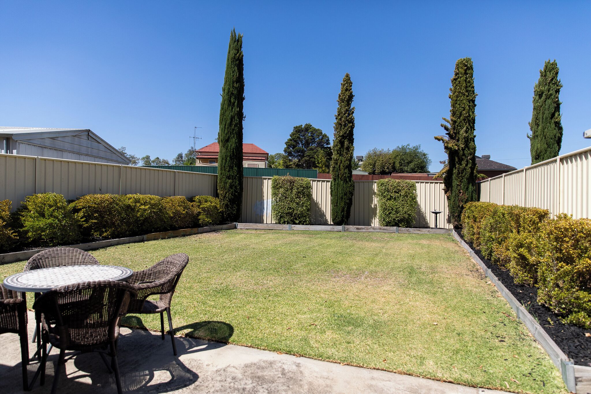 A backyard with a round table and chairs, surrounded by hedges and tall trees.