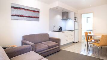 A modern living room with a sofa, a dining area with chairs, a kitchen with a microwave and refrigerator, and a large abstract painting on the wall.