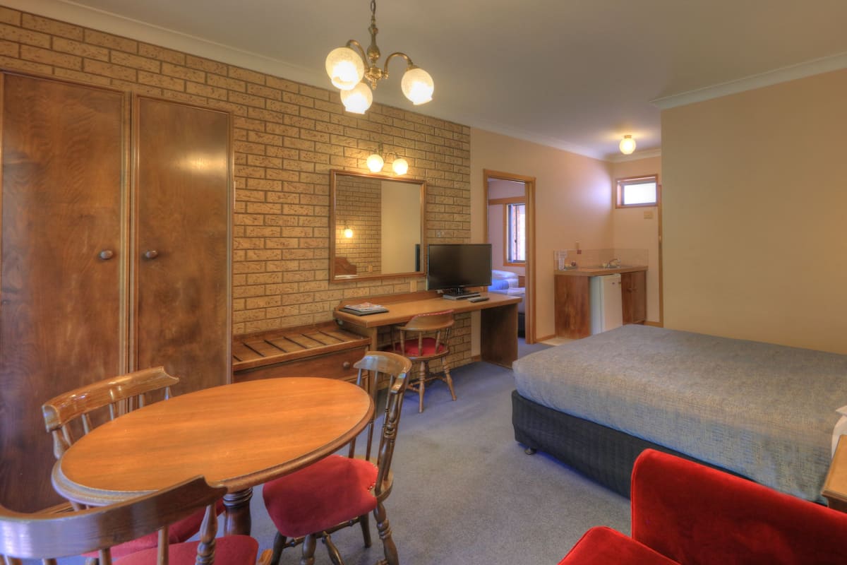 A hotel room with a brick wall, a wooden desk, a television, a bed, and a dining area with chairs.