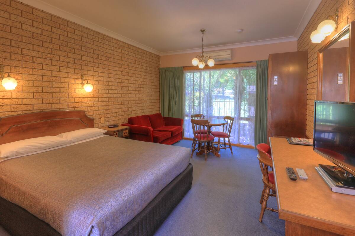 A hotel room with a large bed, a desk with a TV, a red chair, and a dining table with chairs.
