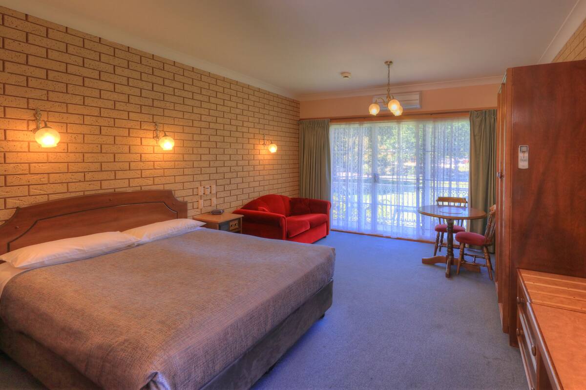 A hotel room with a bed, a red chair, a table with chairs, and a wooden cabinet.
