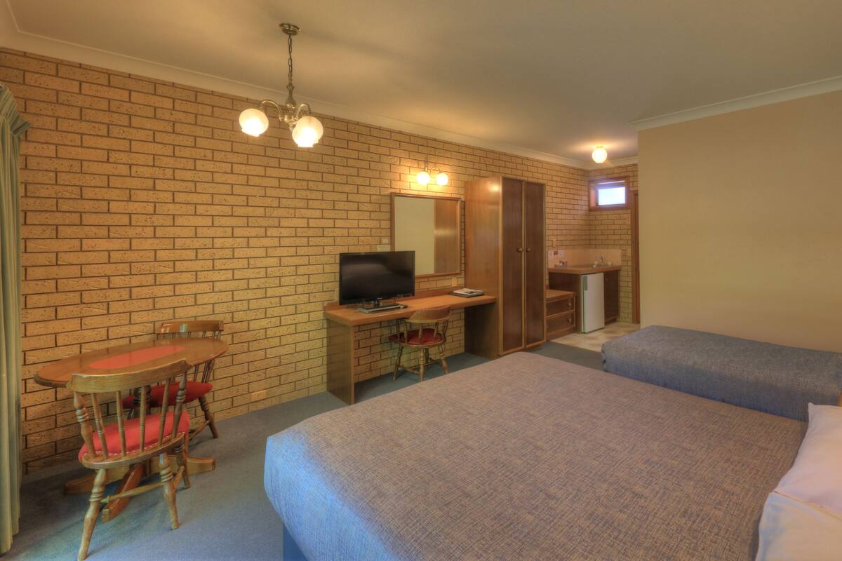 A hotel room with two beds, a desk with a TV, a brick wall, and a kitchenette.