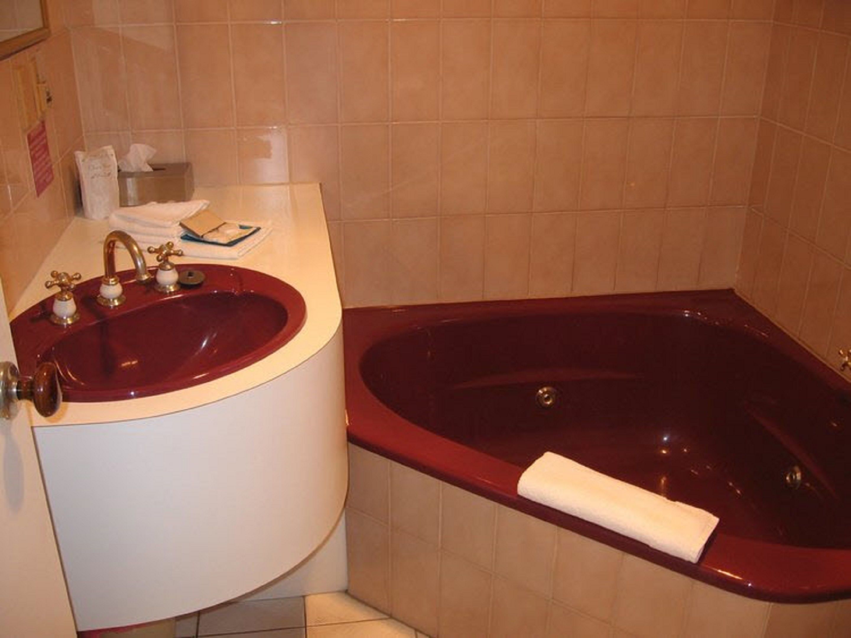 Ein Badezimmer mit einer roten Badewanne, einem weißen Waschbecken mit goldfarbenen Armaturen und beige gefliesten Wänden.