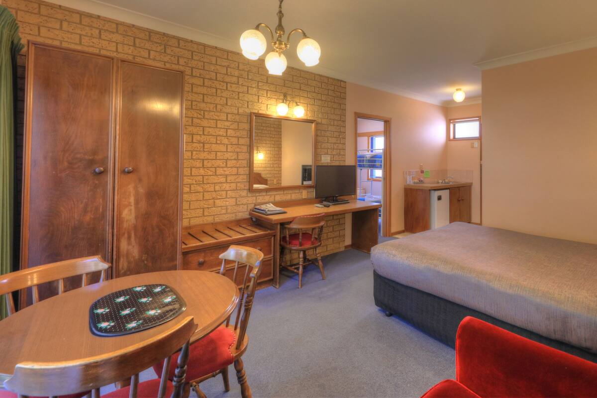 A hotel room with a wooden desk, a chair, a bed, a small kitchenette, and a large mirror.