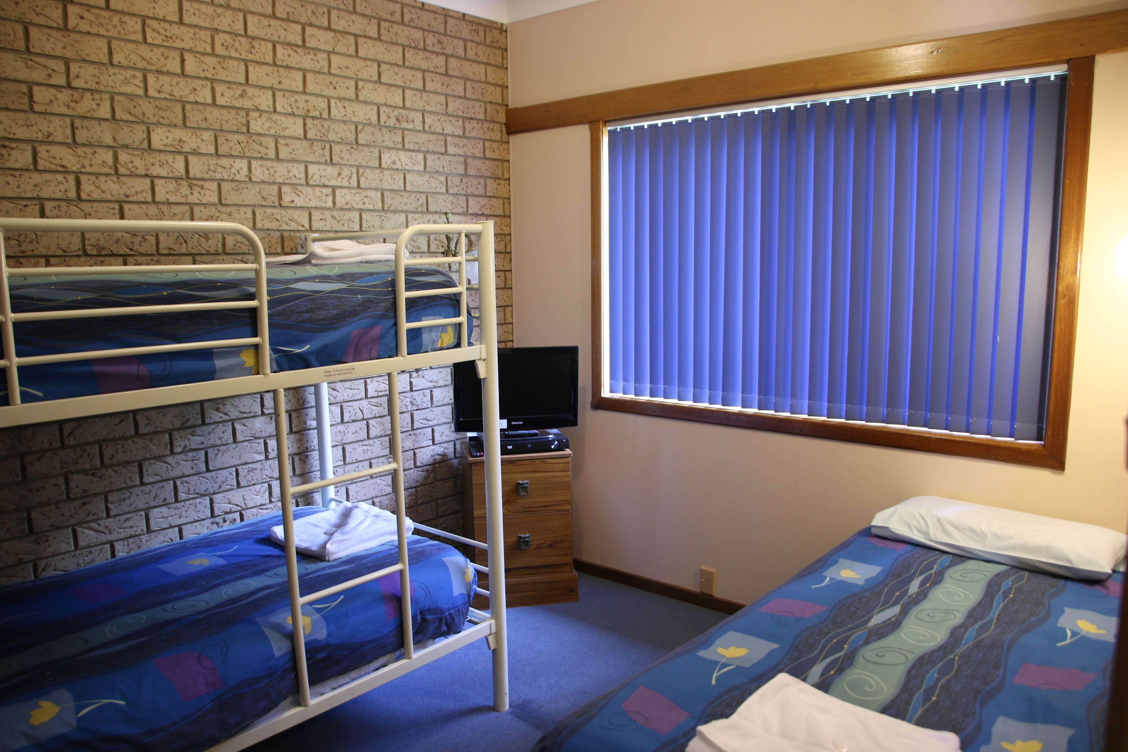 A dormitory room with bunk beds, a television, a window with blinds, and a dresser.
