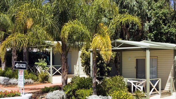 A row of single-story cabins with a covered outdoor seating area, surrounded by a landscaped garden with various shrubs and trees. - Acclaim Kingsway Tourist Park (Madeley)