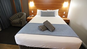Una cama bien tendida con un edredón azul y dos toallas enrolladas, un cabecero de madera, una mesita de noche con una lámpara y una silla a rayas.