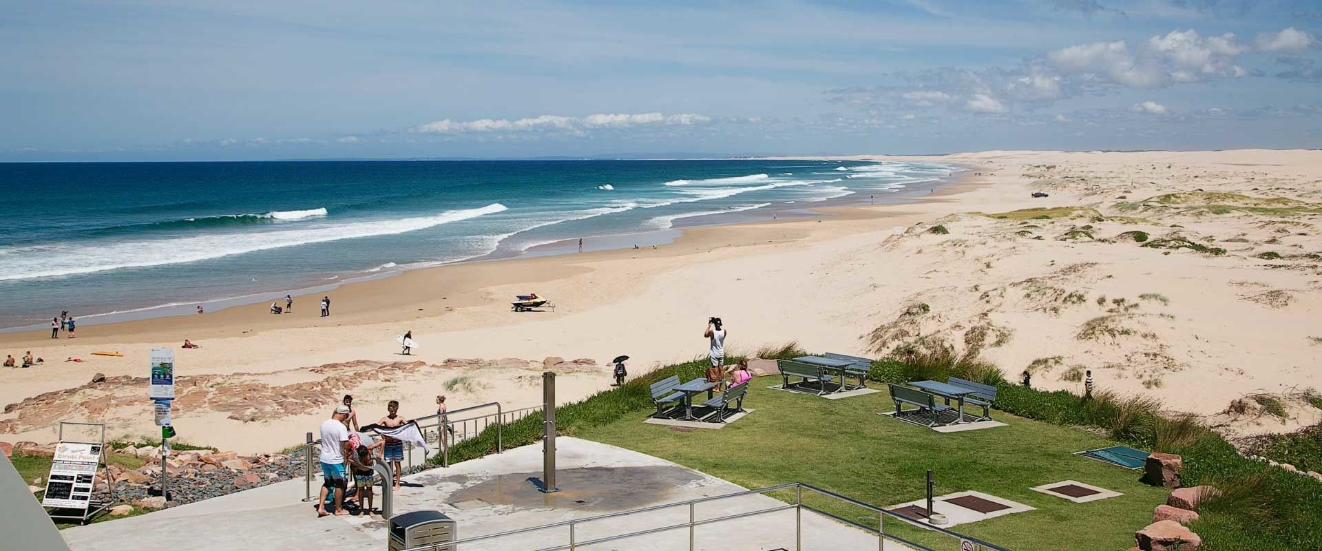 A beach with a sandy shore, a grassy area with benches, and a view of the ocean.